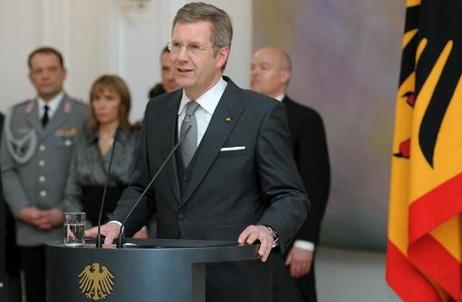 Bundespräsident Christian Wulff steht an einem Rednerpult in Schloss Bellevue. Links von ihm steht die schwarz-rot-goldene Standarte, hinter ihm stehen Männer und Frauen. 