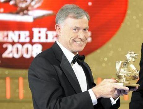 Der Bundespräsident hält die goldene Figur in Form einer Henne in der Hand, im Hintergrund ein Rotes Logo