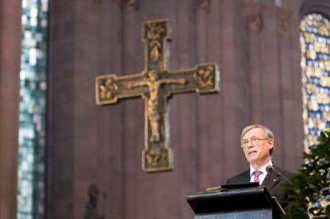 Bundespräsident Horst Köhler spricht von einem Rednerpult. Im Hintergrund sind bunte Kirchenglasfenster zu sehen, der rote Stein des Mainzer Doms und ein christliches Kreuz.