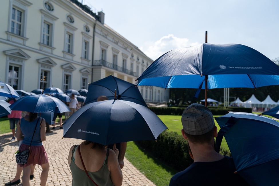 Bürgerinnen und Bürger schützen sich mit Schirmen vor der Sonne während sie beim Bürgerfest 2019 auf einen Rundgang durch das Schloss Bellevue warten 