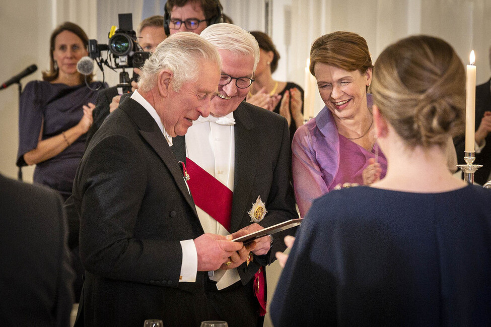Bundespräsident Steinmeier, König Charles III und Elke Büdenbender stehen beieinander und blicken gemeinsam auf ein Kindheitsfoto des Königs.