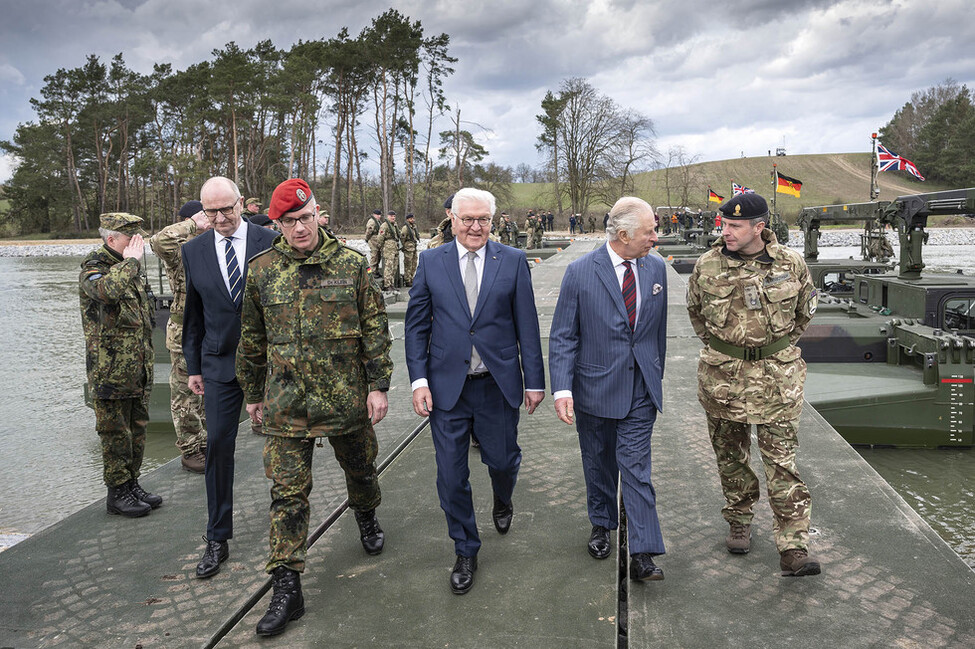 Bundespräsident Steinmeier und König Charles III. gehen über eine Brücke, begleitet von einem britischen und einem deutschen Soldaten.