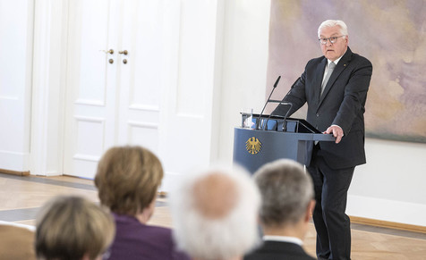 Bundespräsident Steinmeier hält eine Rede an einem Stehpult und schaut die im Publikum sitzende Angela Merkel an.