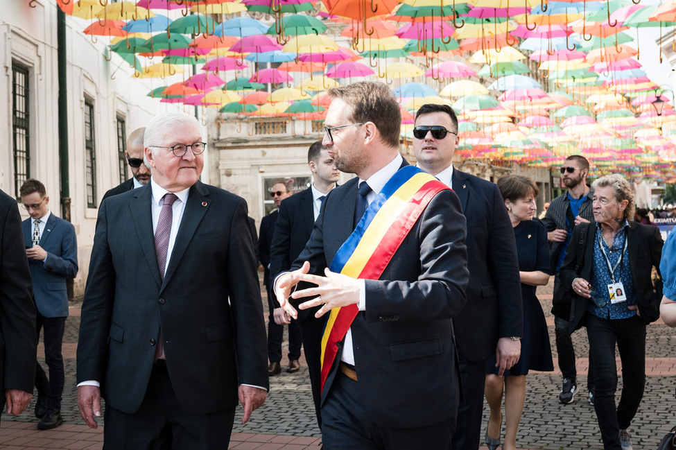 Bundespräsident Steinmeier und Bürgermeister Dominic Fritz laufen auf einer Straße, über ihnen bunte Regenschirme.
