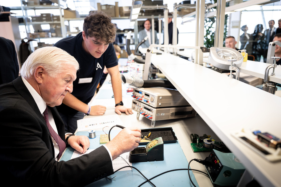 Bundespräsident Frank-Walter Steinmeier sitzt an einem Arbeitstisch in der Ausbildungswerkstatt und lötet zusammen mit einem Auszubildenden ein Bauelement für eine Schaltung während seines Besuchs bei der Firma Adtran (© Bundesregierung/Sandra Steins) Bundespräsident Frank-Walter Steinmeier sitzt an einem Arbeitstisch in der Ausbildungswerkstatt und lötet zusammen mit einem Auszubildenden ein Bauelement für eine Schaltung während seines Besuchs bei der Firma Adtran
