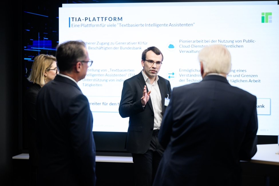 Janek Blankenburg (Mitte) erläutert Bundespräsident Steinmeier und anderen die Einsatzmöglichkeiten für Künstliche Intelligenz anhand einer auf Leinwand projezierten Präsentation