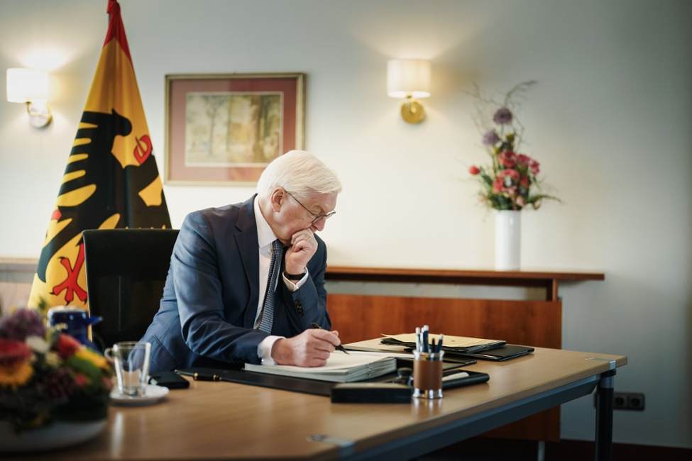 Bundespräsident Steinmeier sitzt an seinem zeitweiligen Amtssitz vor der präsidialen Standarte am Schreibtisch und schreibt in einer Akte (© Bundesregierung/Denzel) Bundespräsident Steinmeier sitzt an seinem zeitweiligen Amtssitz vor der präsidialen Standarte am Schreibtisch und schreibt in einer Akte